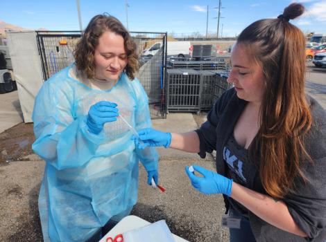 Two people outside wearing protective gear doing a medical procedure with a tube and swqb